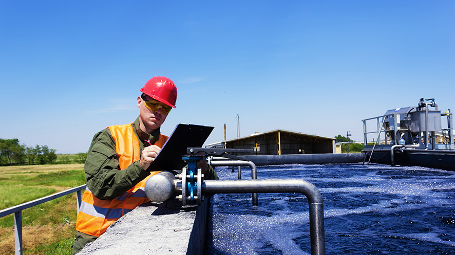 A man in an orange vest and hard hat writing using a clipboard beside an open tank