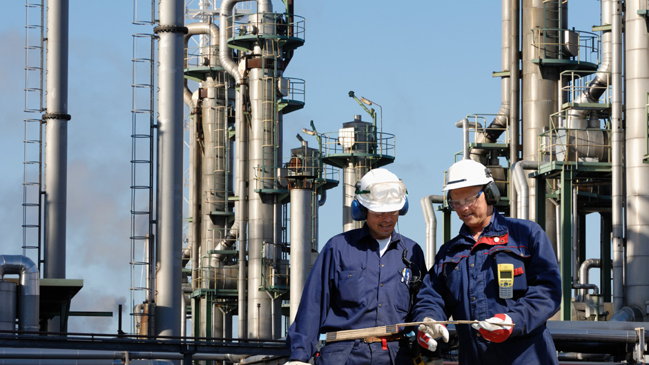 Two men in blue overalls and hard hats standing in front of a refinery