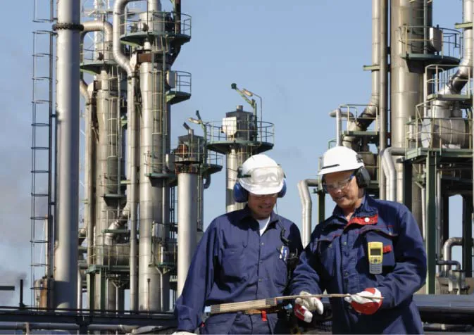 Two men in blue overalls and hard hats standing in front of an oil refinery