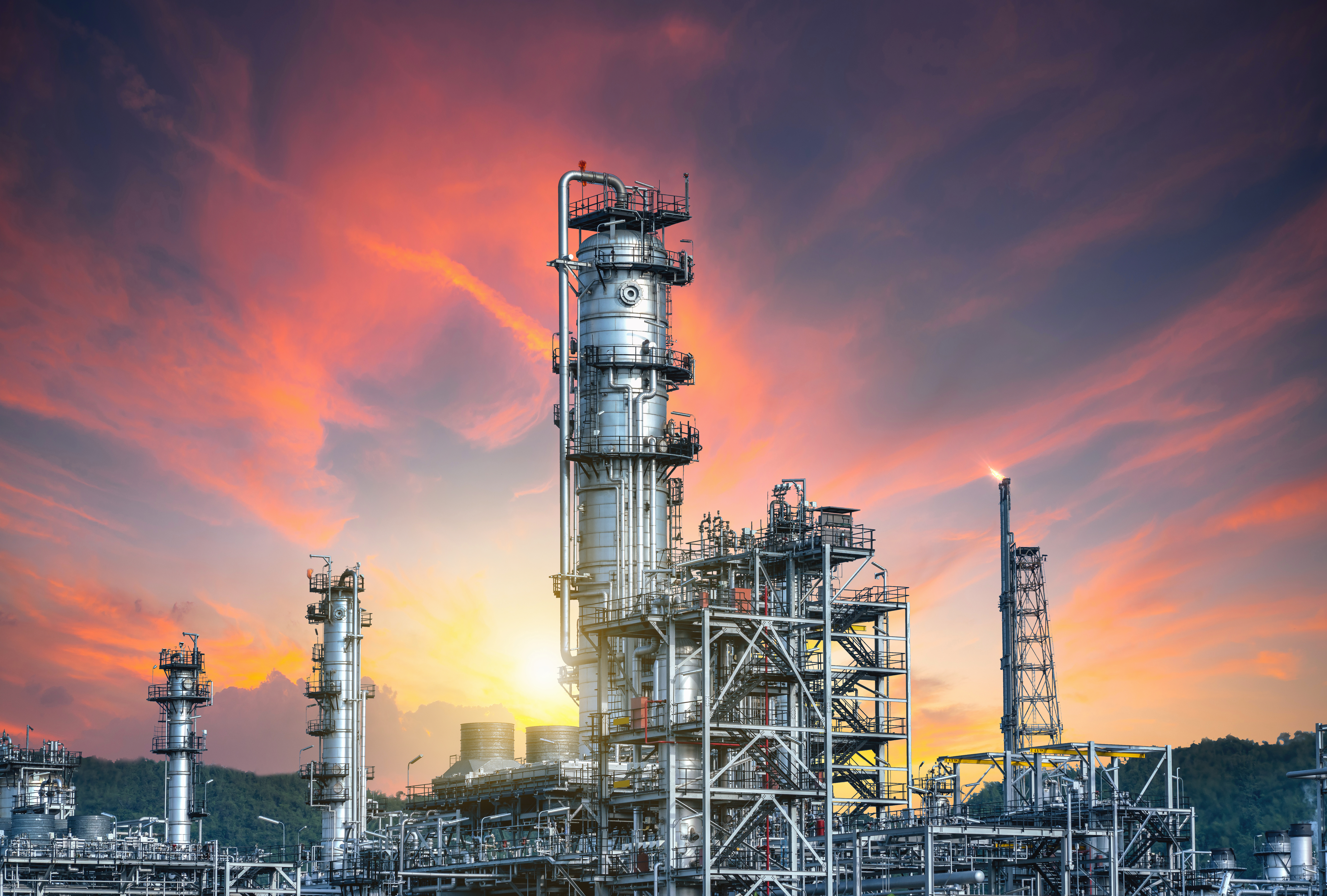 Industrial refinery plant with tall metal distillation towers and pipework, set against a vivid sunset sky with orange, pink, and purple hues.