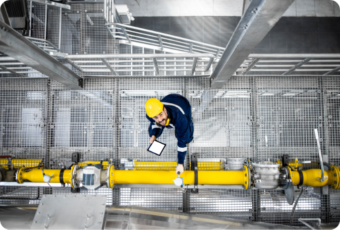 A person in a hard hat and safety glasses stands on a metal walkway with a hand on a yellow pipe.